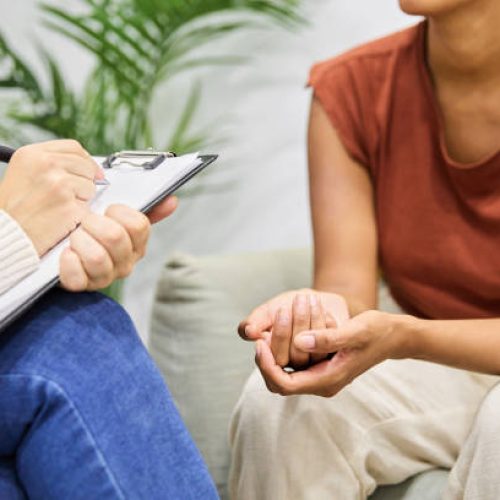 Faceless professional psychologist taking notes on clipboard while counseling woman on couch in office